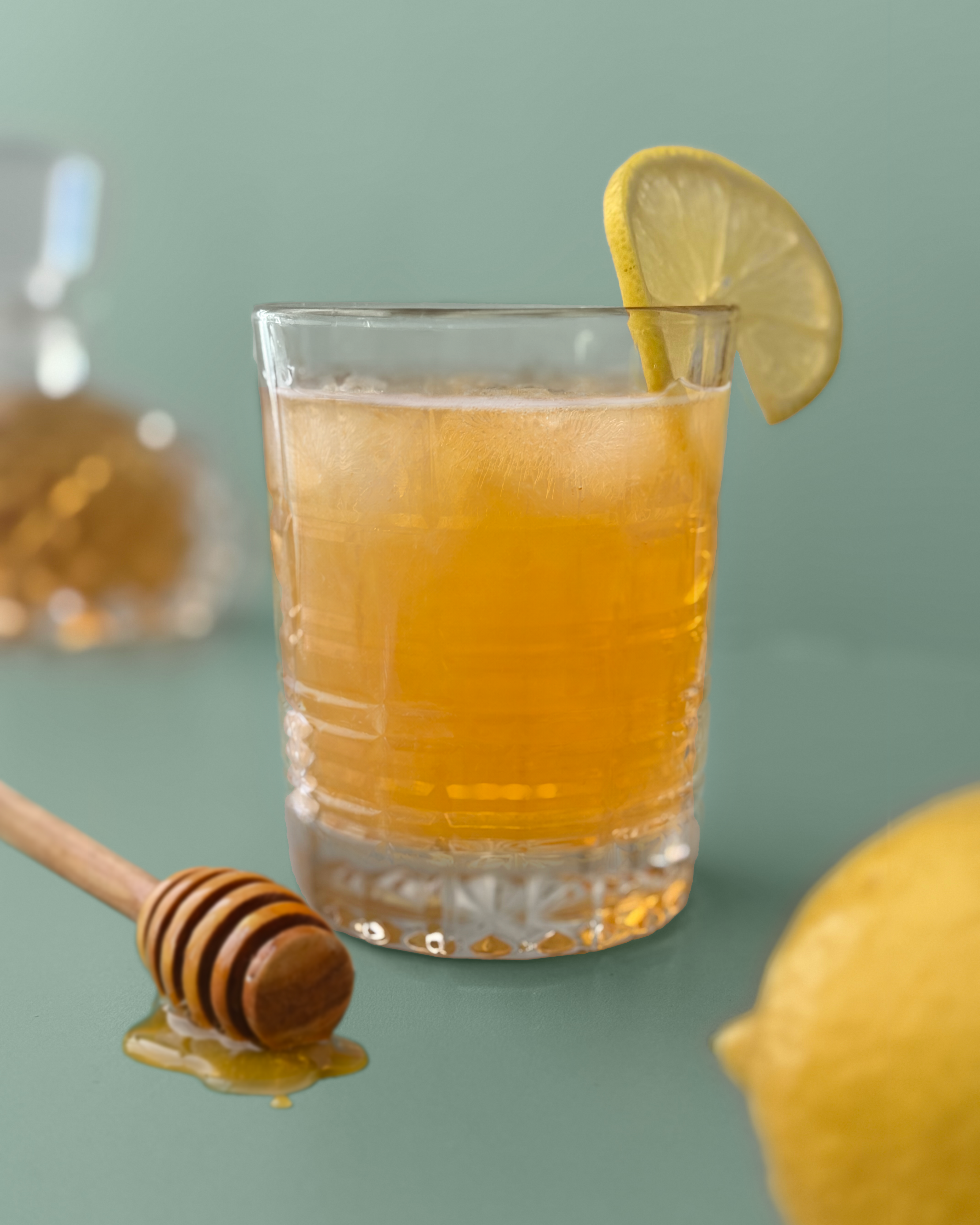 Glass or Irish Gold Rush cocktail with a slice of lemon garnish on the rim of the glass whiskey bottle in the background and a lemon and honey dipper in the foreground on a green background.